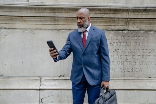 Senior businessman in a blue suit using a smartphone while holding a bag, standing against a stone wall.