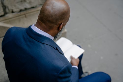 Free Rear view of a man in a suit reading a book outside on a city sidewalk. Stock Photo