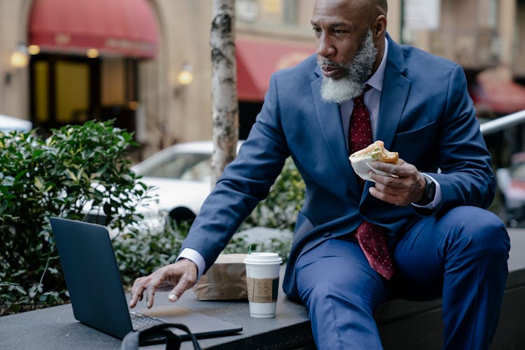 A Businessman Holding A Sandwich While Using A Laptop