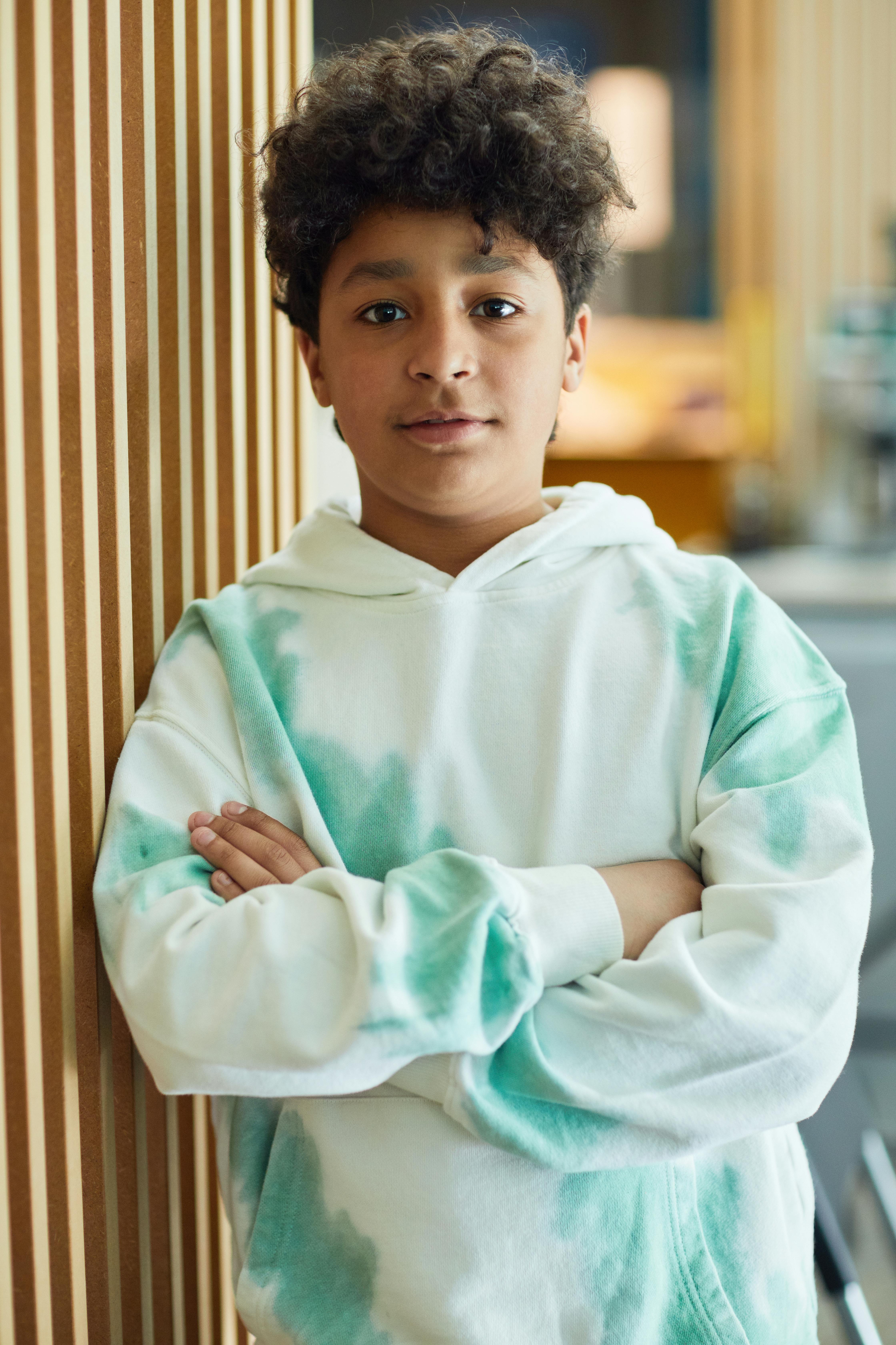Portrait of a young boy with crossed arms wearing a white and green tie-dye hoodie, posing confidently indoors.