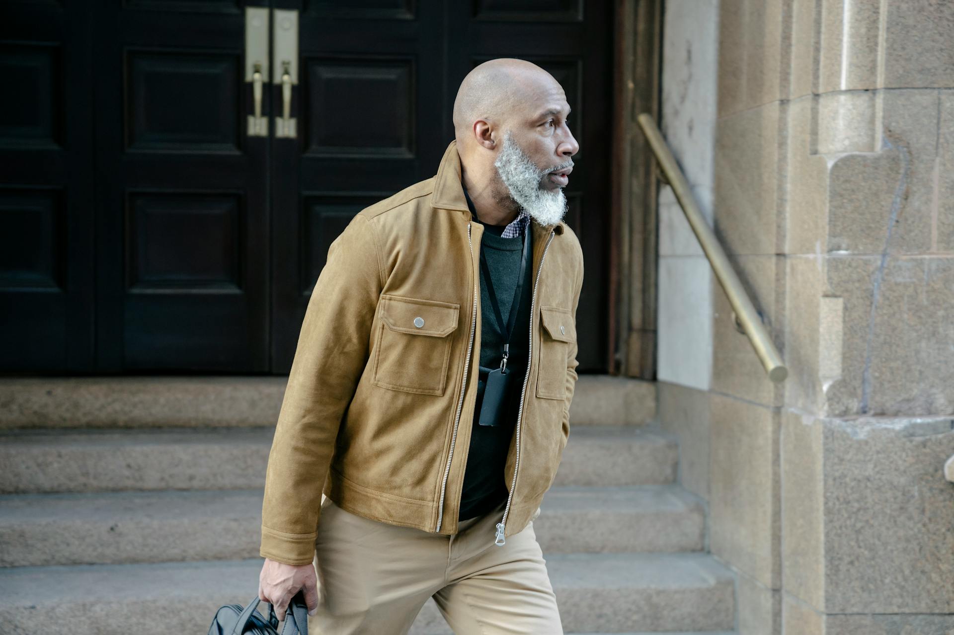 Portrait of a businessman with a beard wearing a stylish jacket, holding a briefcase, and exiting a building.