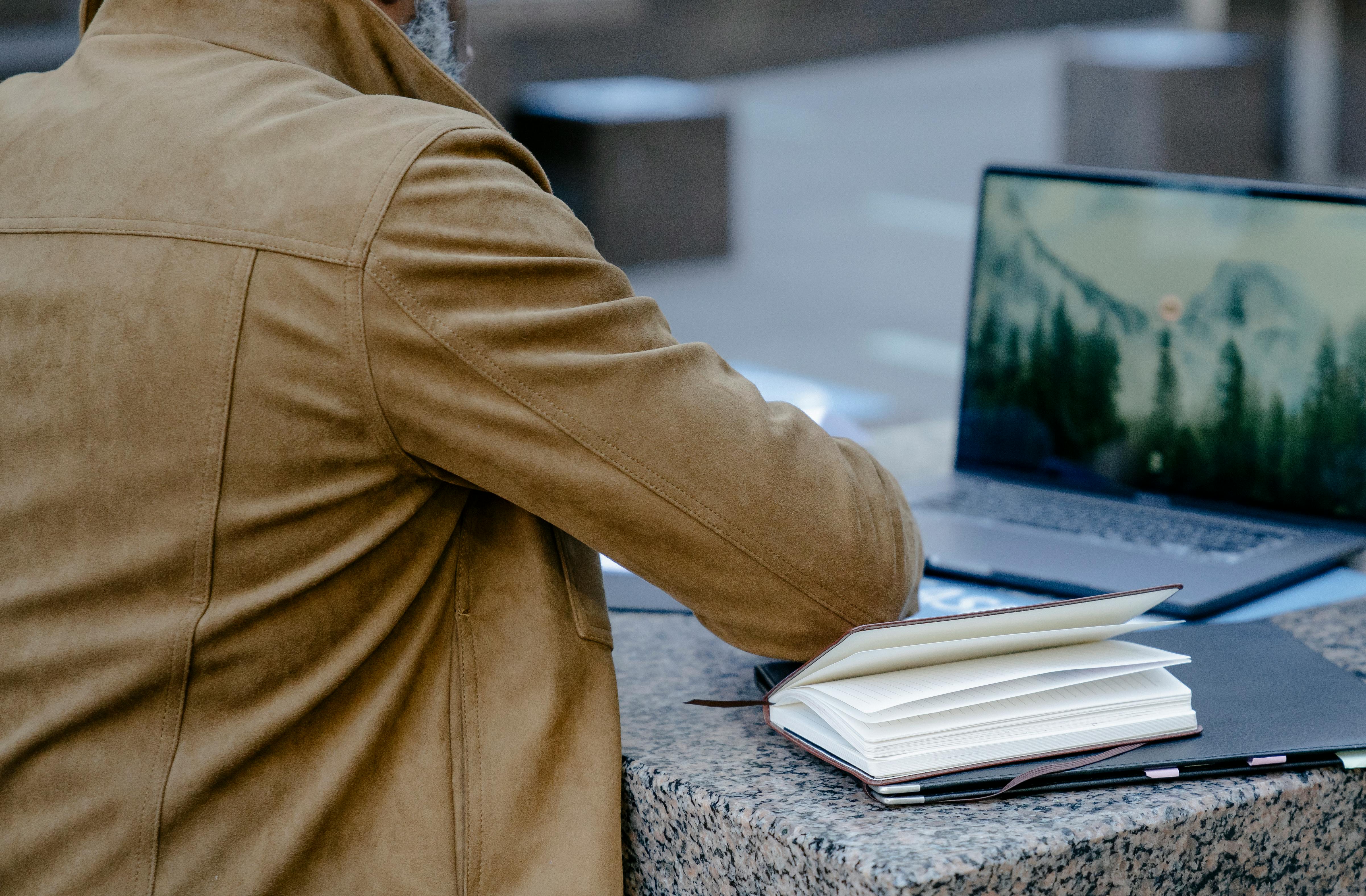 Back view of a businessman working on a laptop and notebook outdoors in an urban setting.