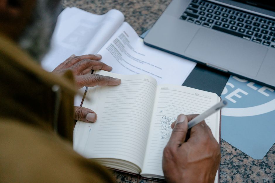 Businessman writing in notebook at desk with laptop and documents.