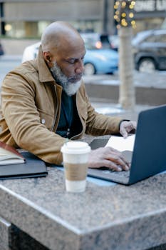 Bald man in a brown jacket using a laptop outdoors with a coffee cup on a marble table.