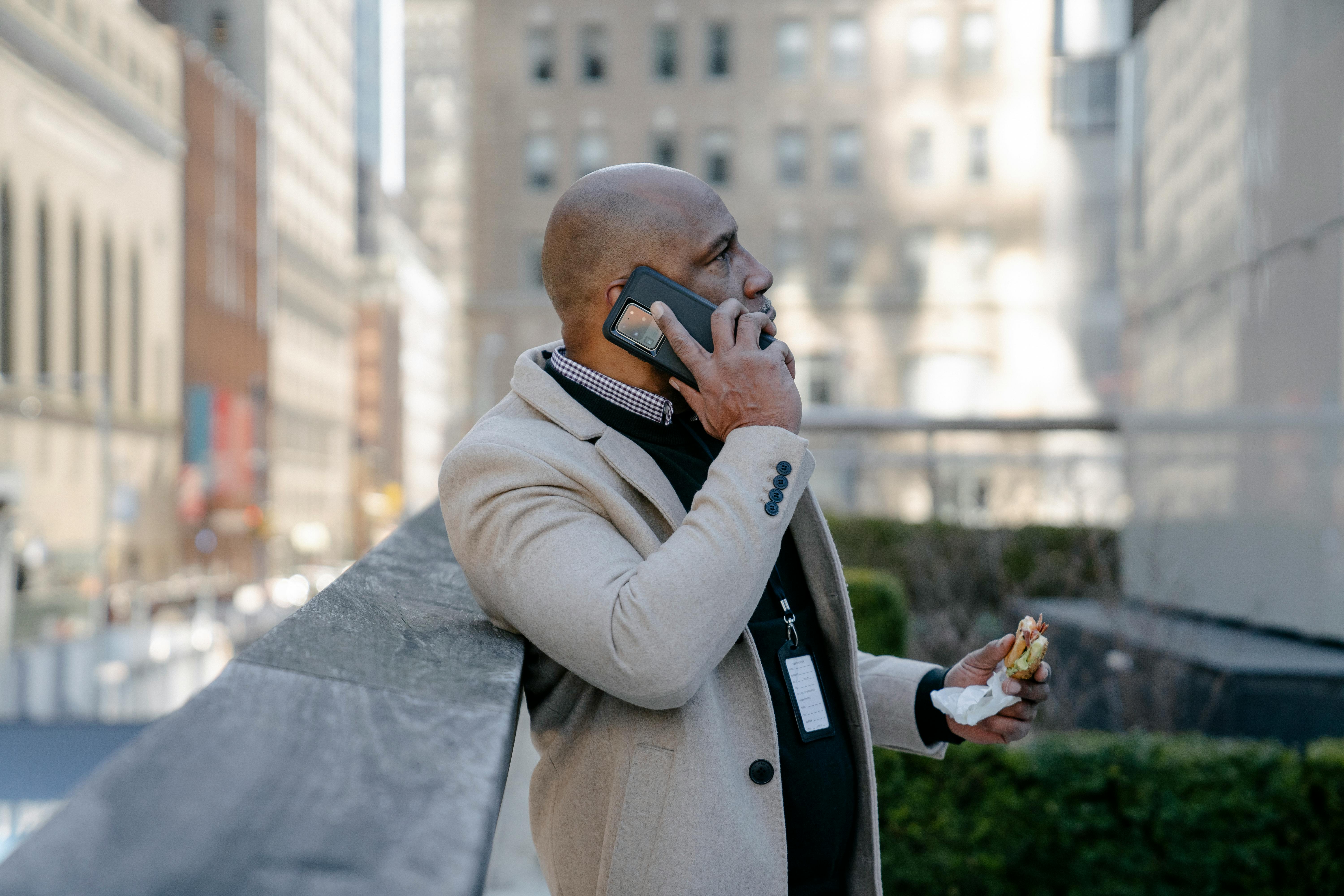 Man in formal attire talks on phone while enjoying a sandwich outdoors in a city setting.