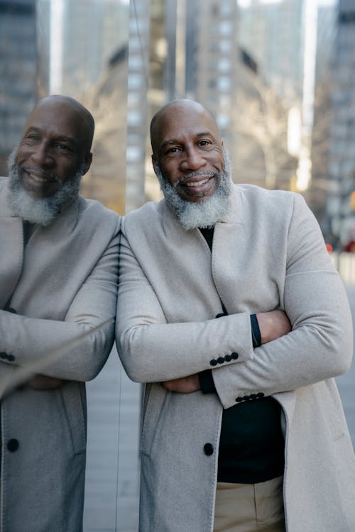 Mature Man with a Gray Beard Elegantly Dressed Leaning Against a Glass Wall of a Building in City 