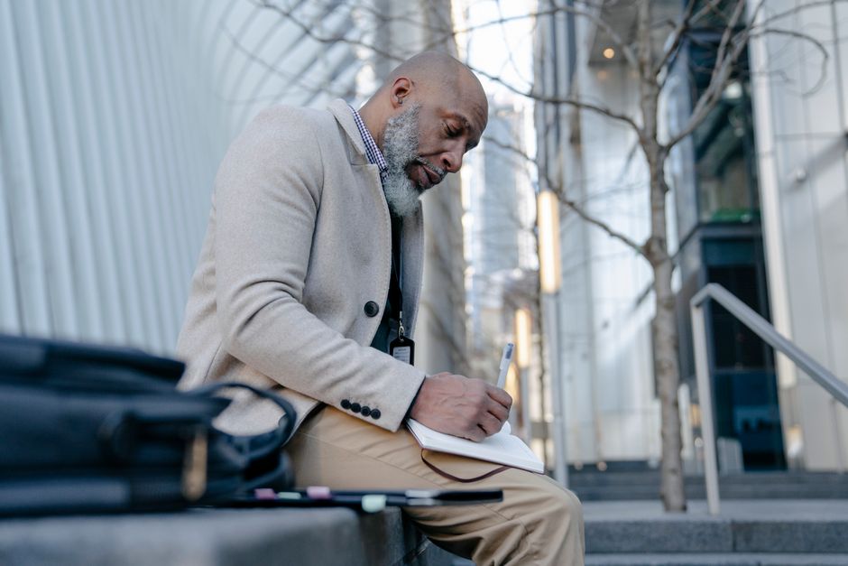 An elderly man in a suit sits outdoors writing in a notebook, capturing a moment of focus in an urban setting.