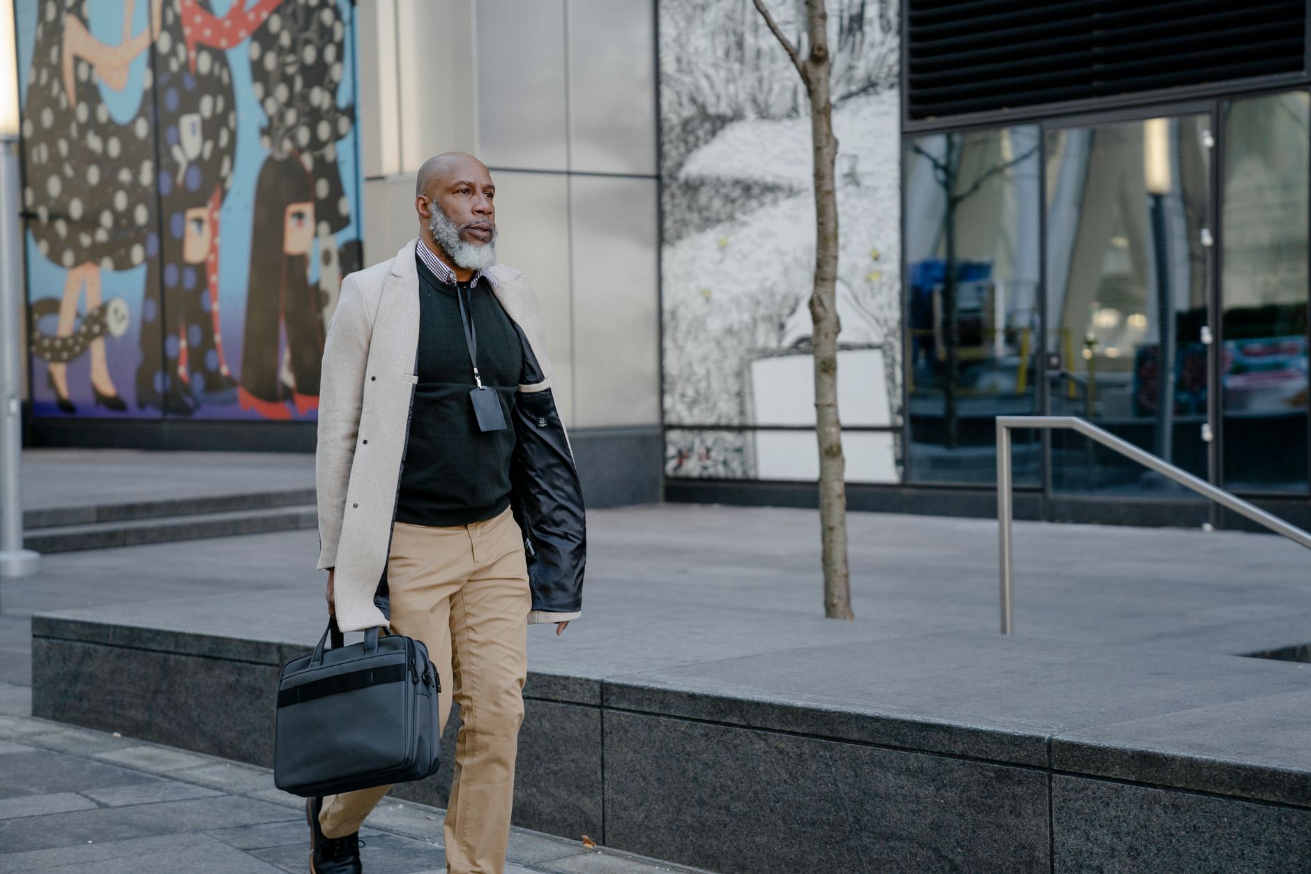 Stylish man with beard carrying a briefcase on a city sidewalk with mural art in the background.