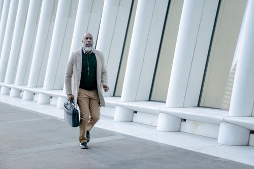 Free Senior man in a tan coat walks along a contemporary building facade, carrying a briefcase. Stock Photo