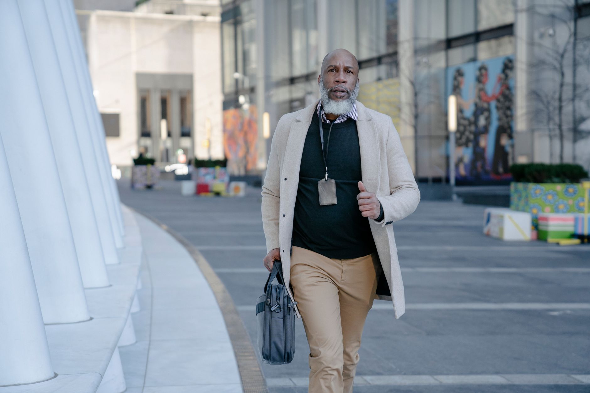 Elderly man with a beard walking outdoors holding a handbag, showcasing urban style.