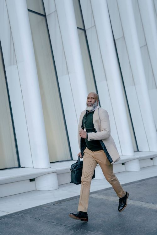 Free Senior man in beige coat and brown pants walking along modern urban architecture carrying a bag. Stock Photo