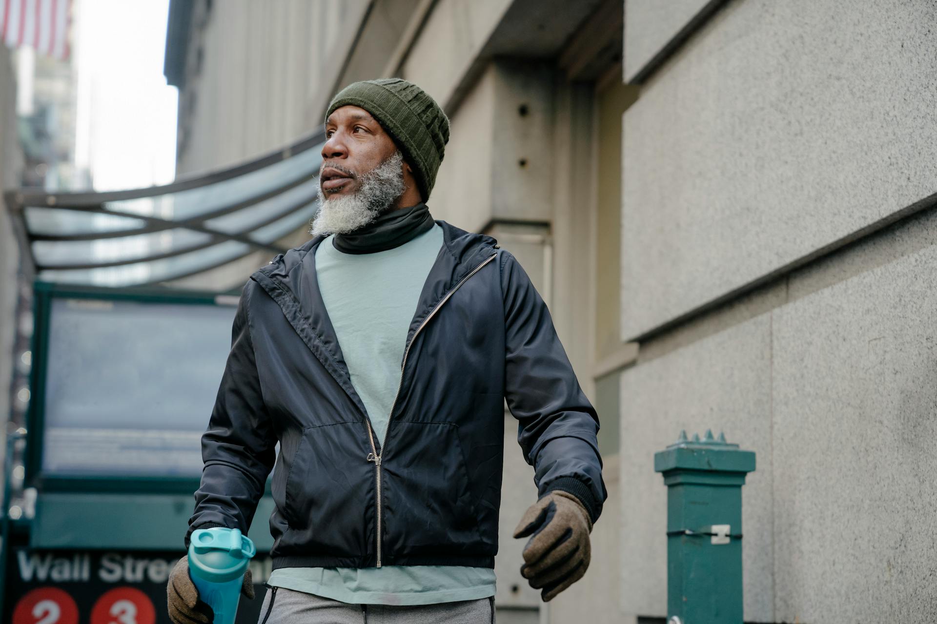 A mature man in active wear walks near a Wall Street subway entrance, carrying a water bottle.