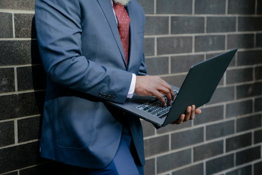 Mid-adult businessman in blue suit typing on laptop against brick wall outdoors.