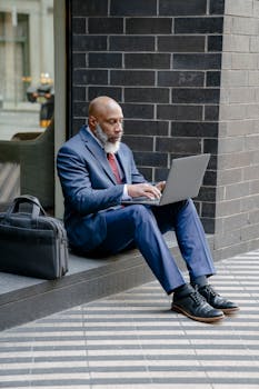 Adult man in suit sitting outside with laptop, symbolizing remote work lifestyle.