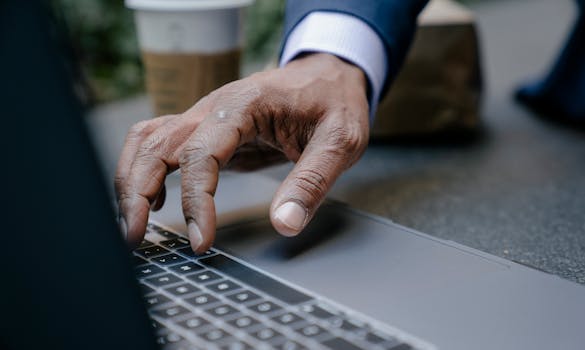 A close-up of a man's hand typing on a laptop keyboard outdoors.