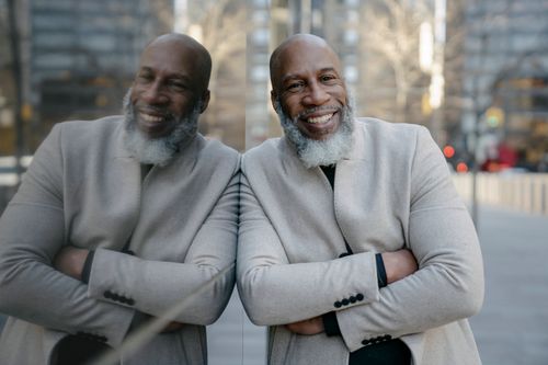 Free An African American man with a beard smiles confidently, leaning against a reflective glass wall. Stock Photo