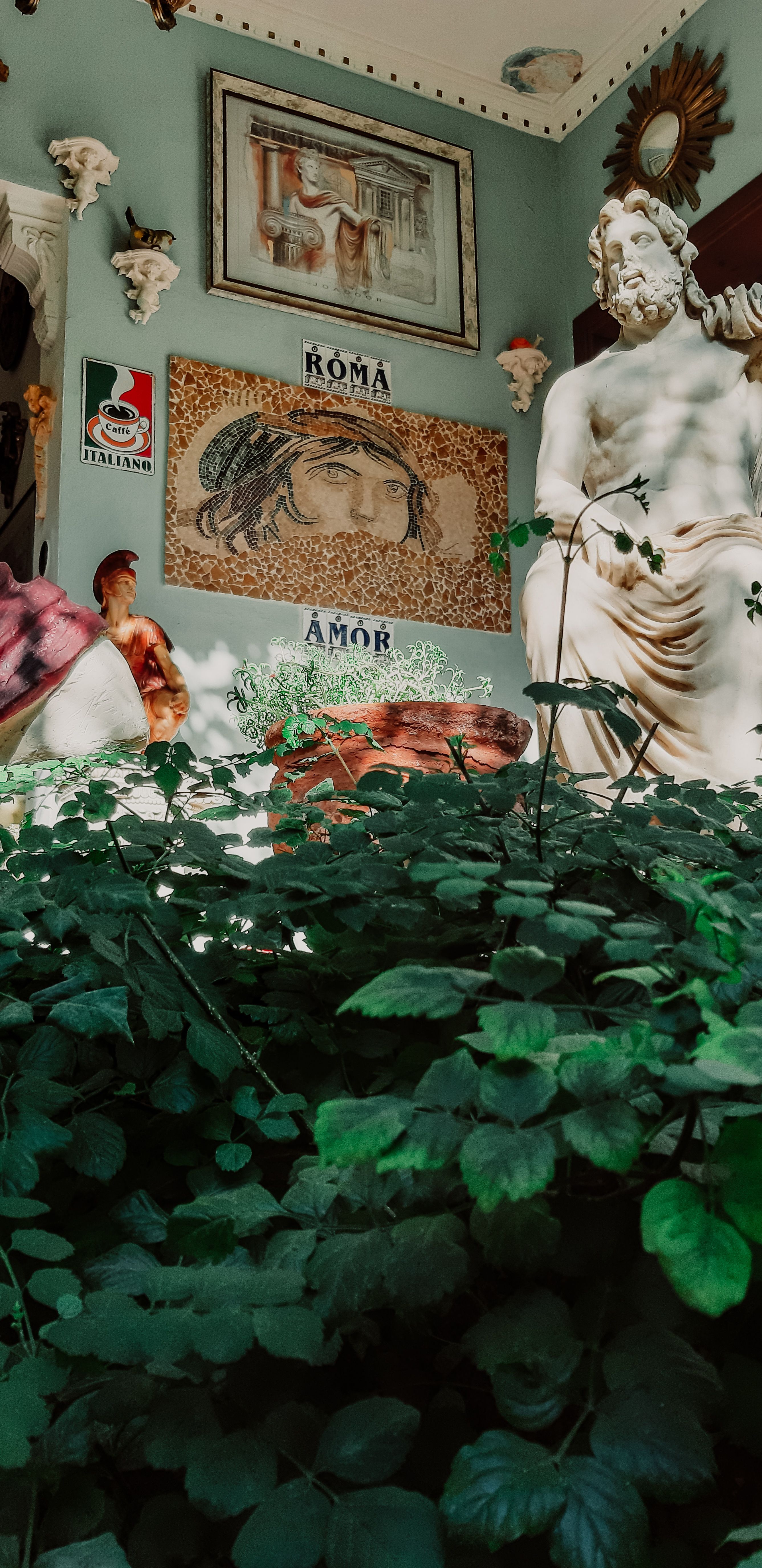 A decorative wall with art pieces and statues amidst lush green plants in Muğla, Türkiye.