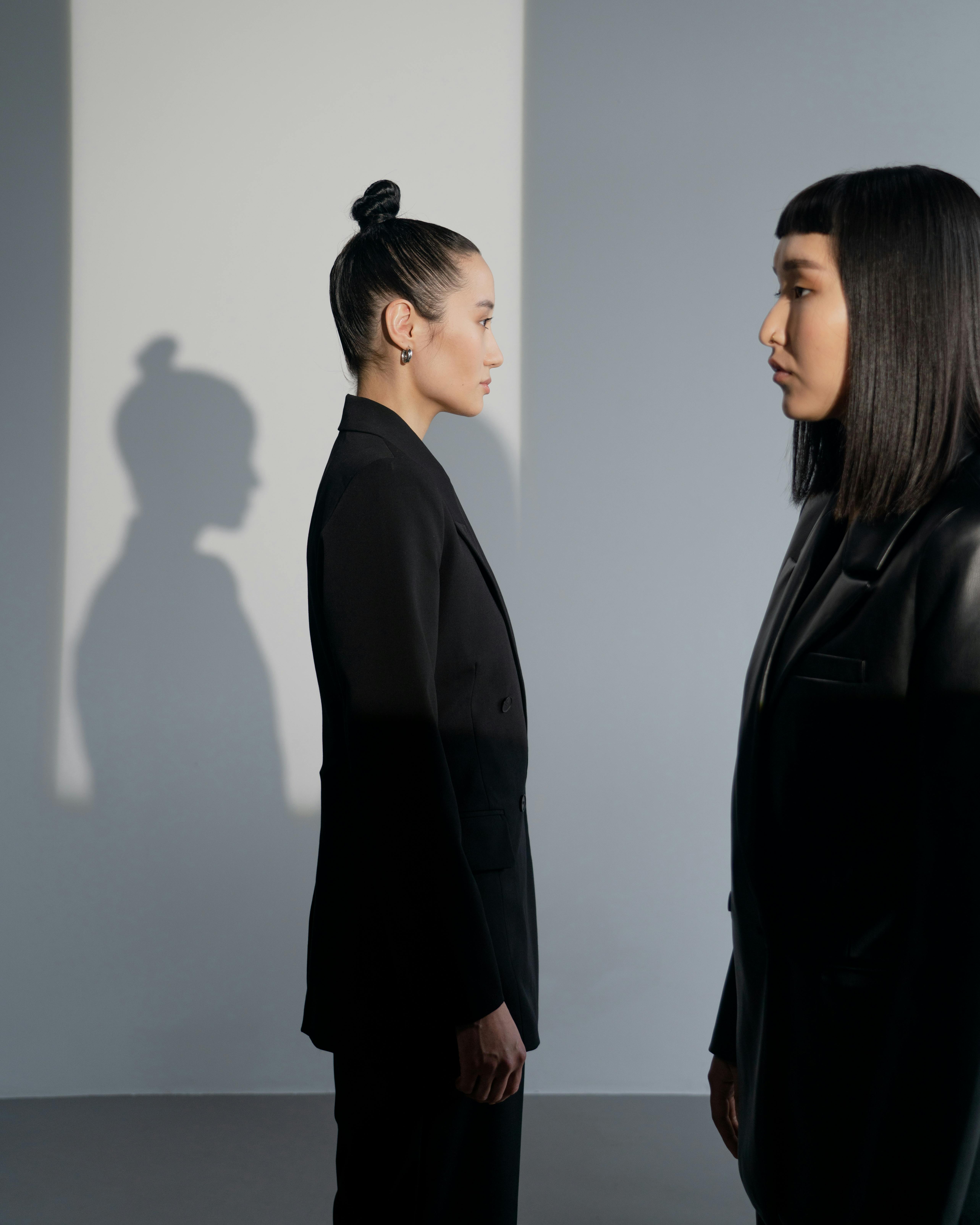 Two women in stylish black outfits stand face-to-face in a well-lit studio, casting unique shadows.