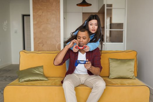 A happy interracial couple playing video games together on a sofa, embracing and having fun indoors.