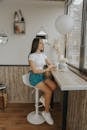 Young contemplative female in gumshoes and denim shorts sitting on chair with takeaway hot drink while looking out window in cafeteria