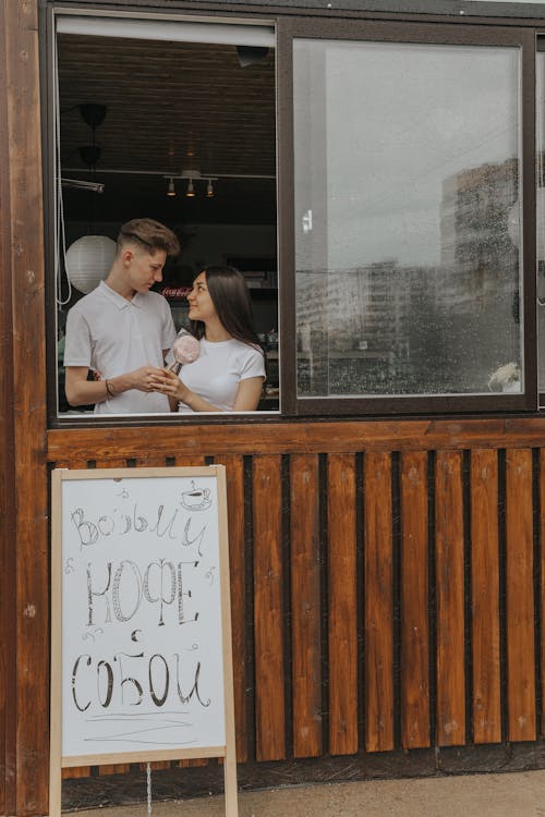 Young man with female beloved looking at each other against cafeteria window and stand with inscription on urban pavement