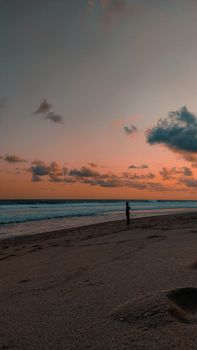 Silhouette of a person on a beach at twilight, under a dramatic sky and calm sea.