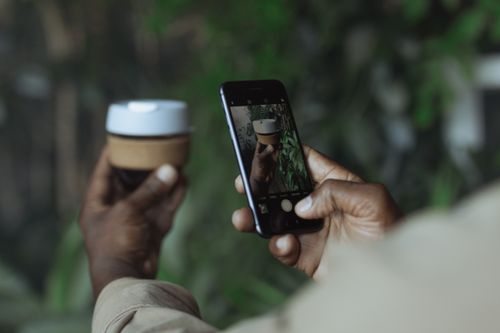 Free An adult uses a smartphone to capture a reusable coffee cup in a blurred green background. Stock Photo
