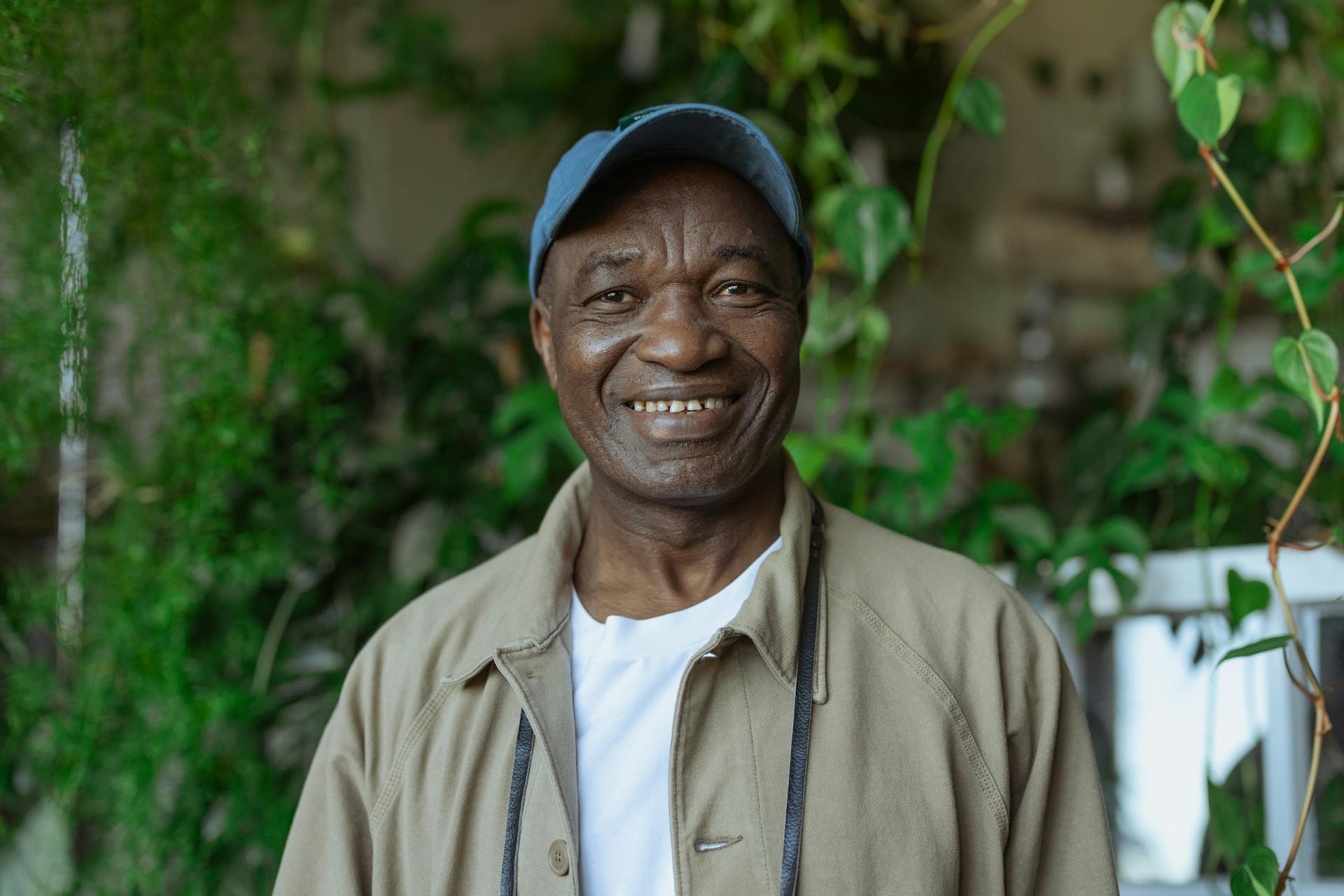 A happy senior man wearing a cap and jacket, standing in front of lush indoor plants.