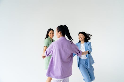 Three young Asian women joyfully holding hands and dancing in trendy pastel outfits against a white studio background.