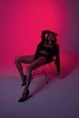 Seductive woman in underwear and jacket sitting on chair with hand behind head while looking at camera in neon light