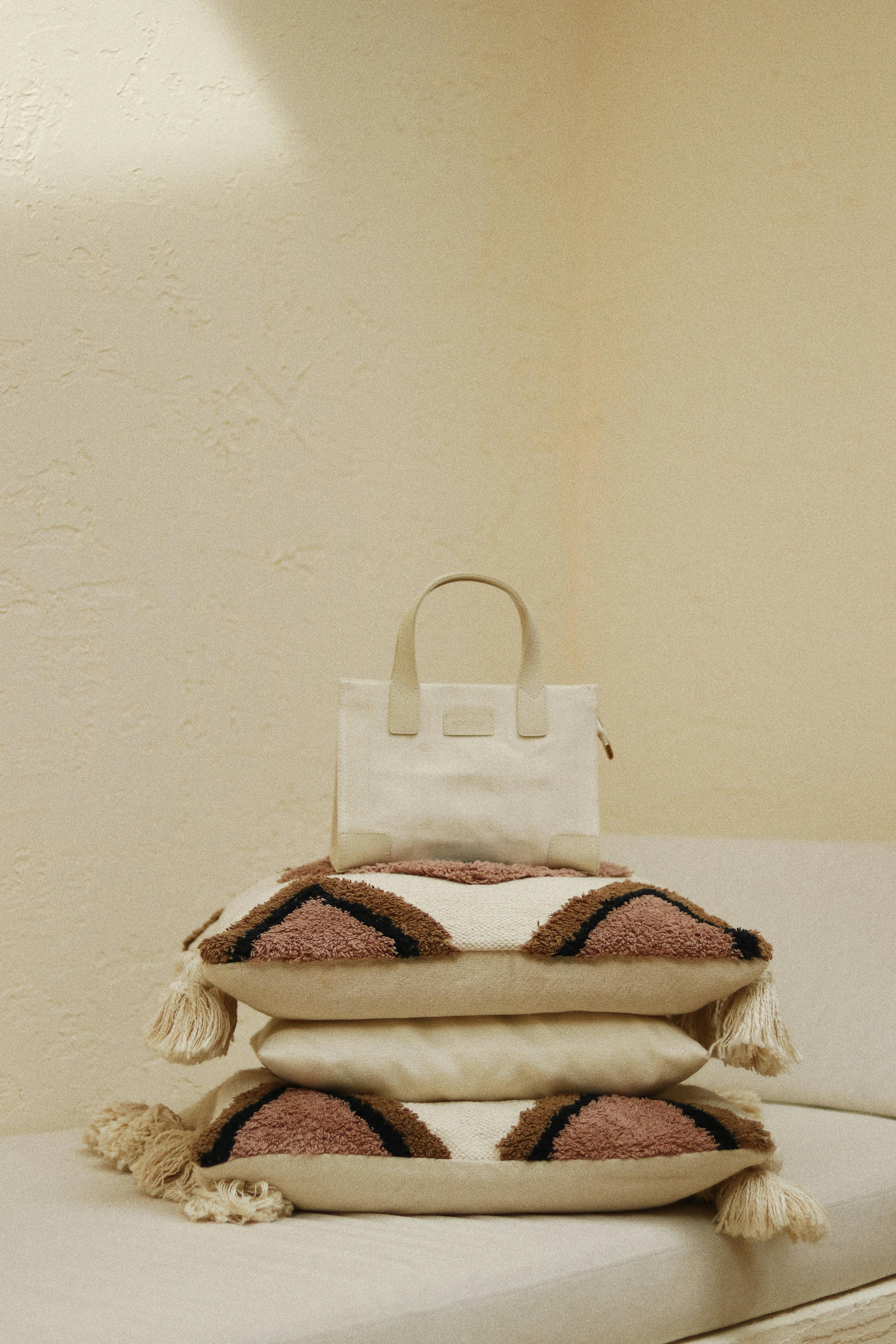 Trendy handbag on ornamental cushions with tassels on textile bench in house on beige background