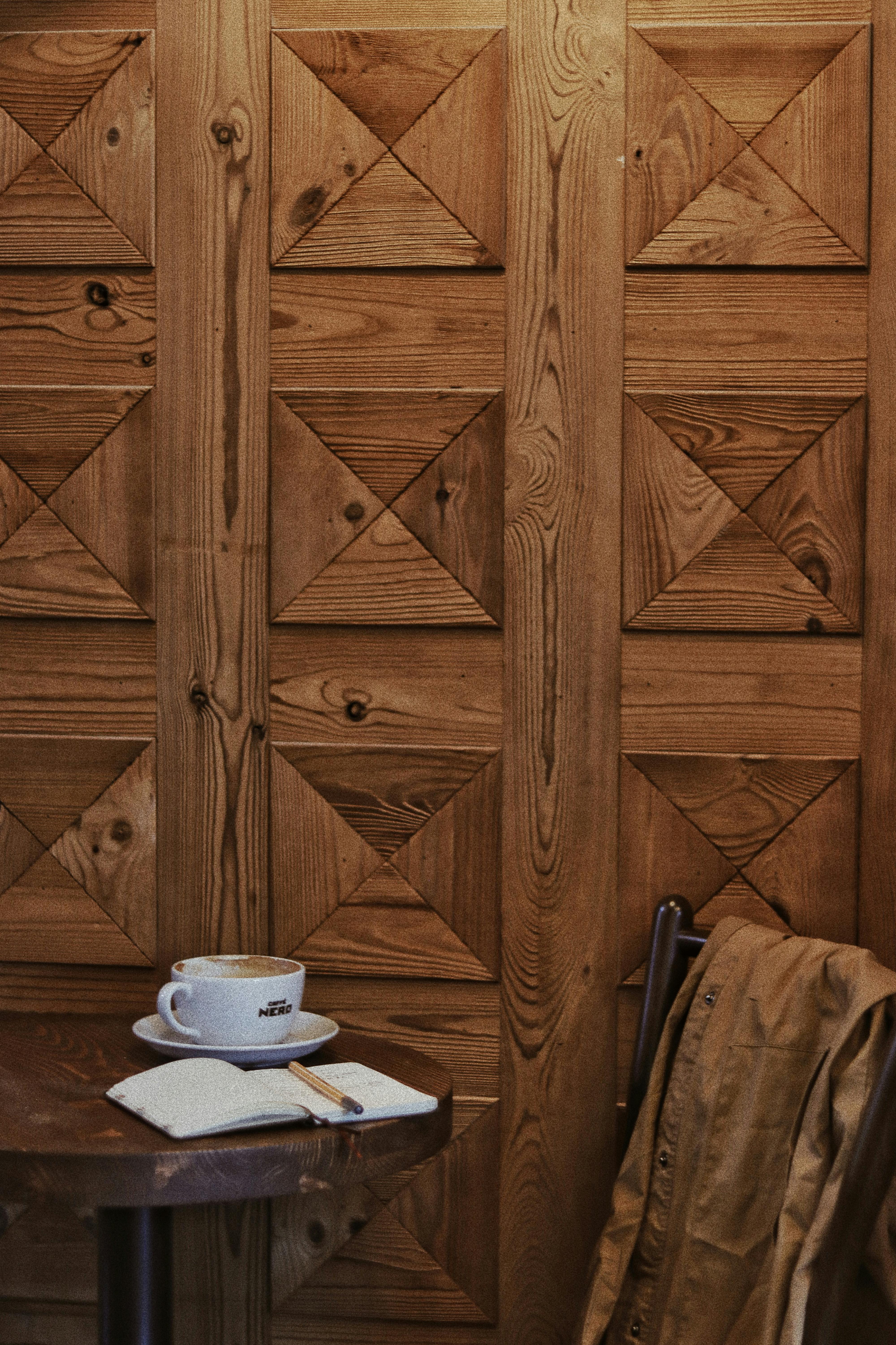 Agenda with pen near ceramic cup of aromatic coffee on table against chair with coat and wooden wall in cafe