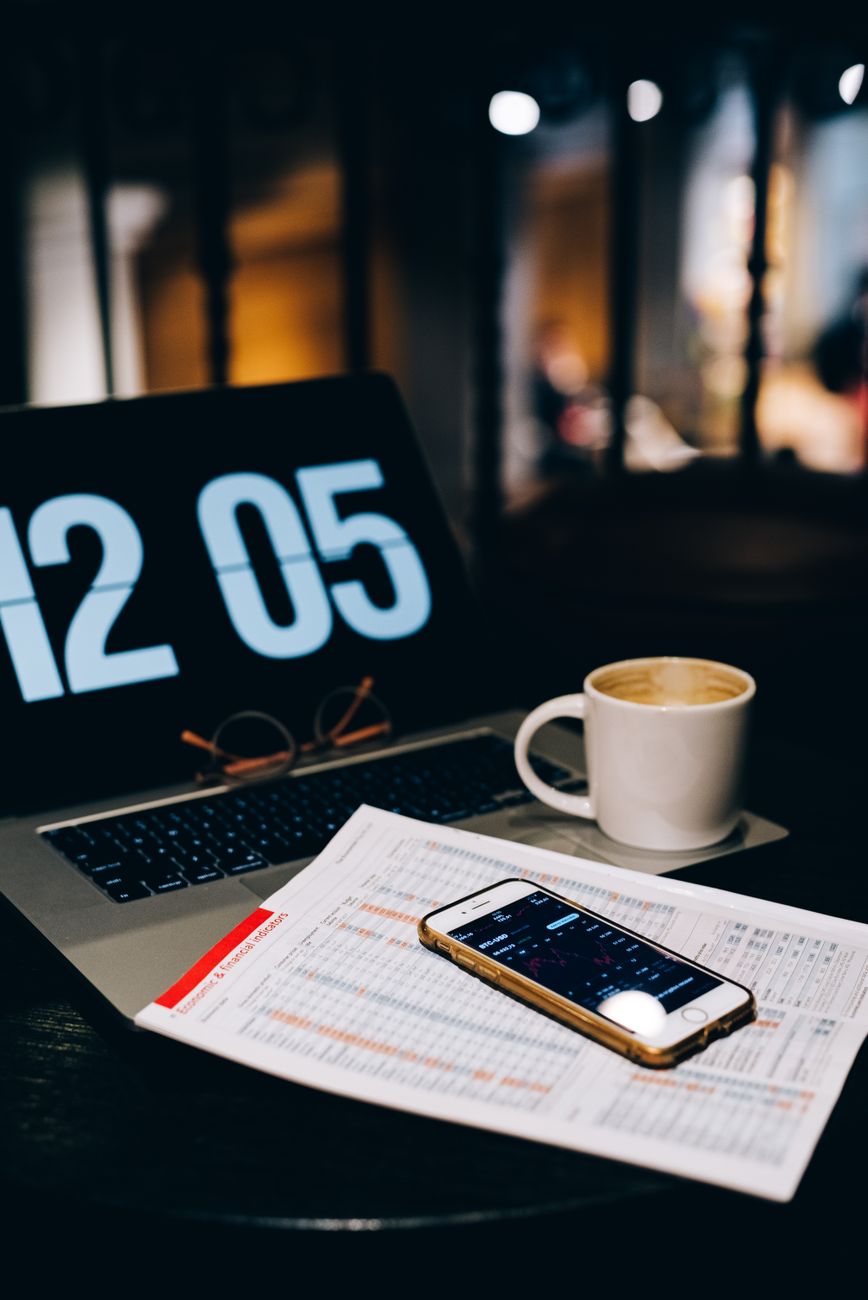 Hand holding a phone with a banking app showing simple spending charts, next to a cup of coffee and a laptop