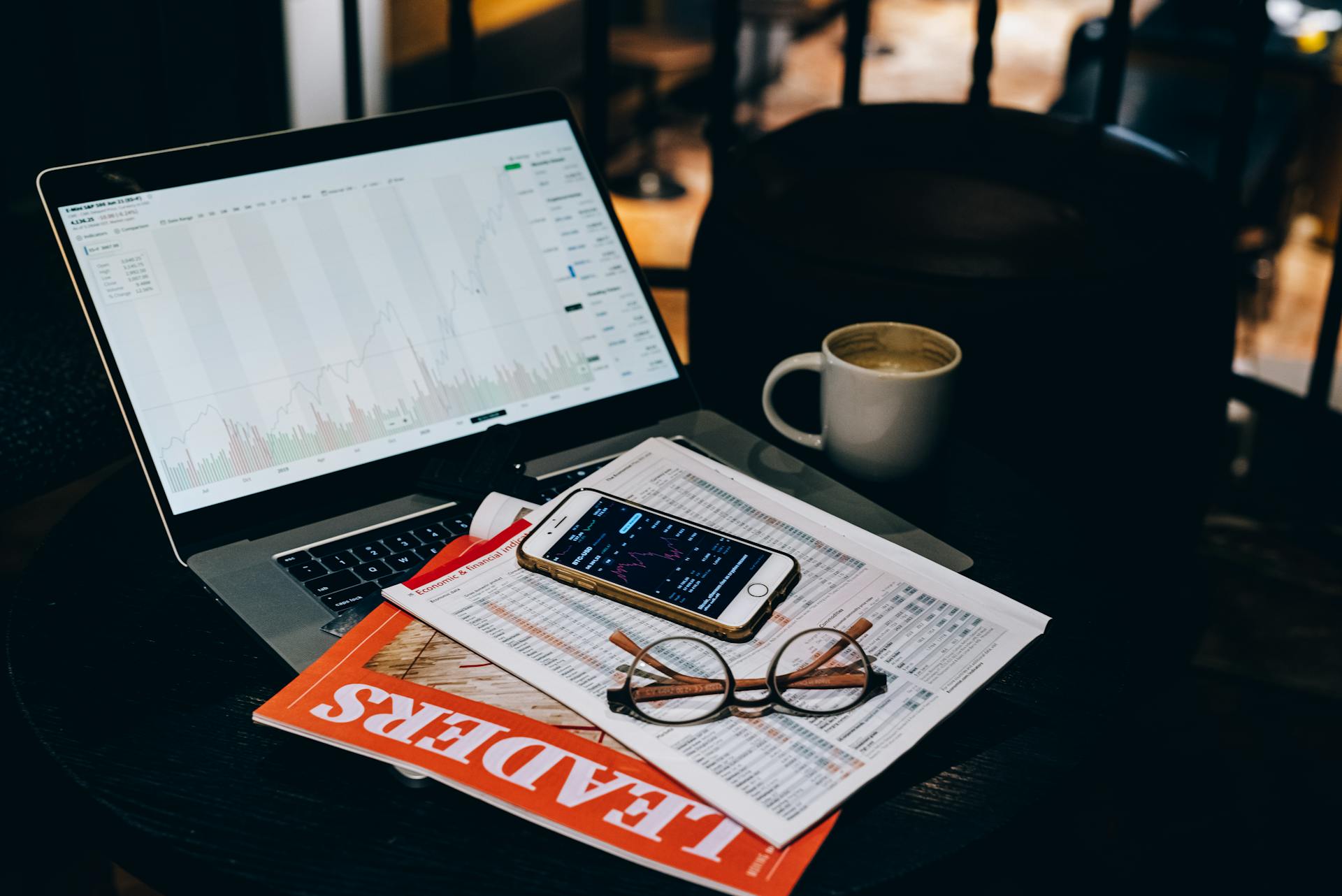 A cozy office desk setup with a laptop displaying stock charts, coffee, and documents.