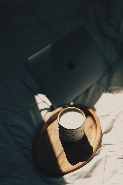 From above of ceramic cup of cappuccino placed on wooden plate near opened contemporary netbook on comfortable bed in sunlight