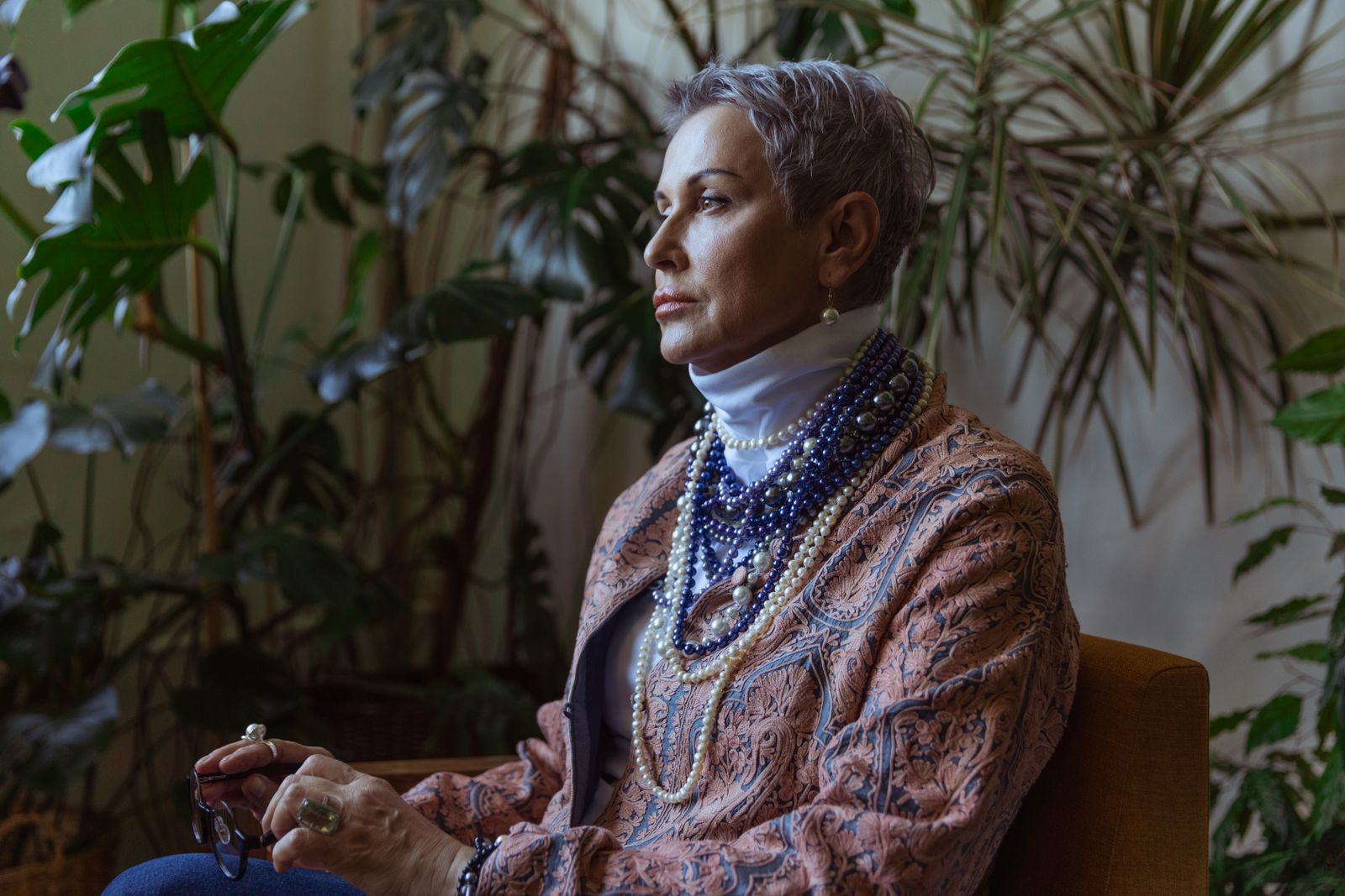Silver-haired woman in patterned jacket and layered pearl necklaces sits calmly among green houseplants