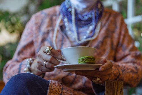 Free A senior woman holding a tea cup with a green macaron, showcasing elegance and bohemian style. Stock Photo