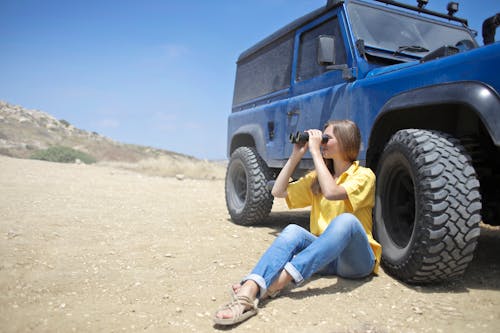 Seorang Wanita Duduk Di Tanah Di Samping Jeep Dengan Menggunakan Teropong