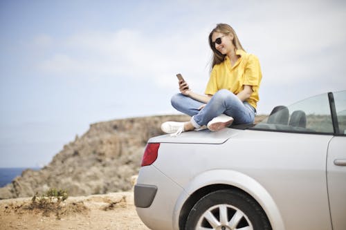 Frau In Der Gelben Bluse Und In Den Blauen Jeans, Die Selfie Beim Sitzen Auf Auto Nehmen