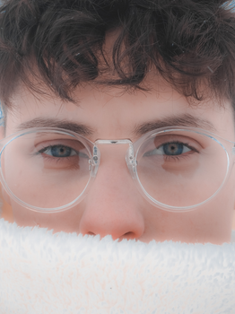 A close-up portrait of a person wearing round clear eyeglasses, partially covered by a white fluffy fabric.