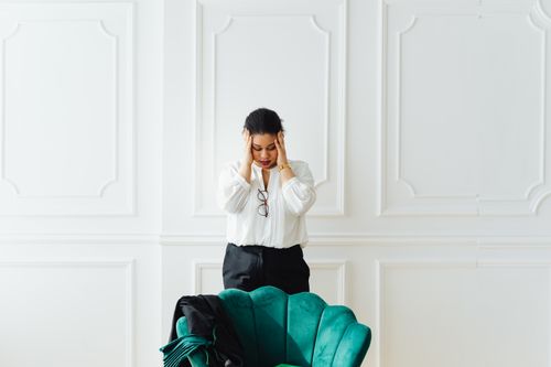 Free A woman holds her head in stress, standing indoors next to a green chair. Stock Photo