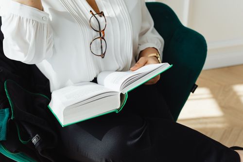 Free Adult woman reading a book indoors, holding glasses. Sunlit scene with a cozy atmosphere. Stock Photo