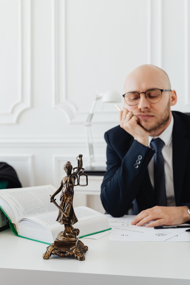 A Statue Of Lady Justice On A Desk Of A Man Wearing Eyeglasses