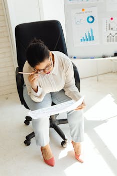 A woman in an office chair reviews finance charts and graphs, holding papers with intense focus.