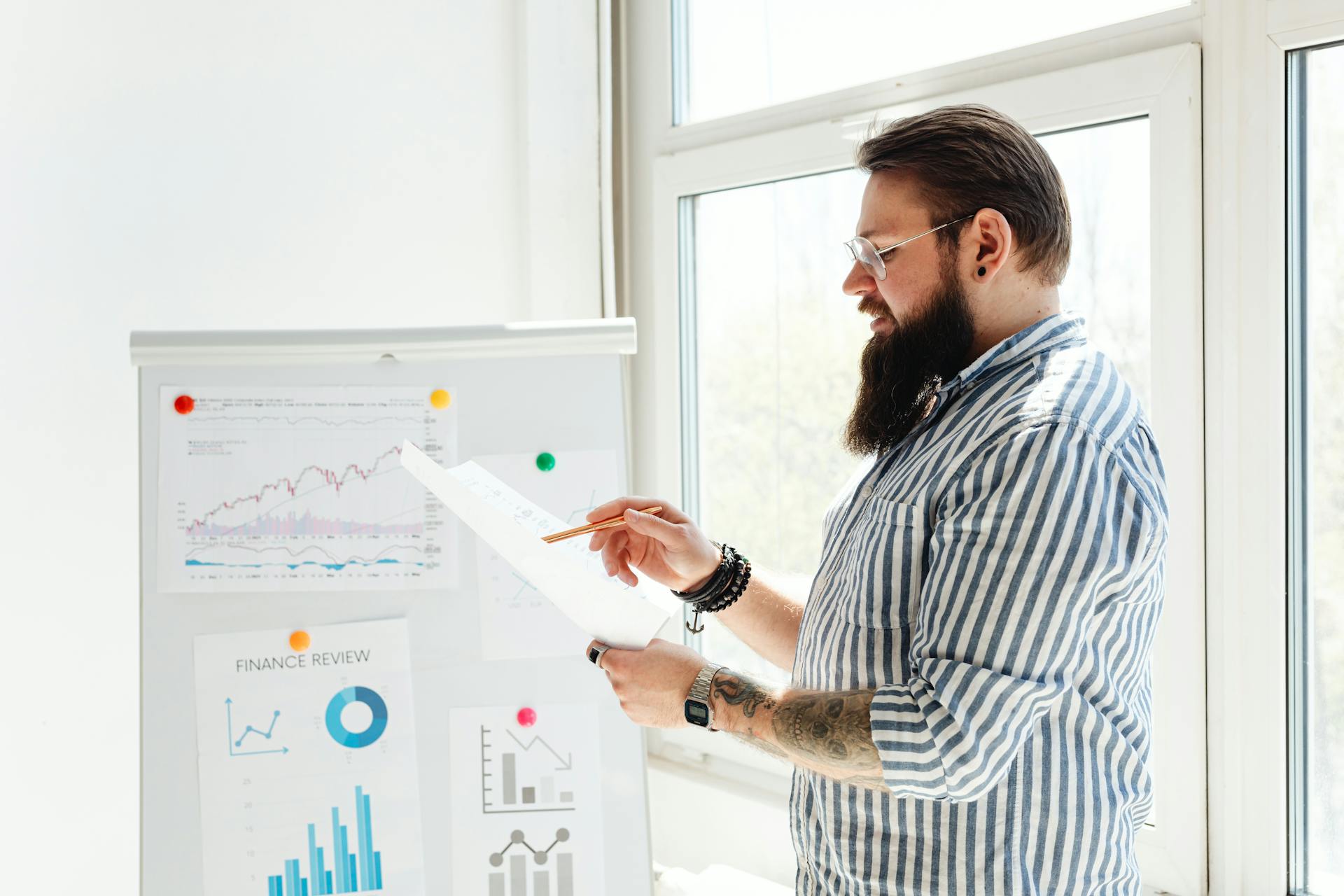 A bearded man in glasses reviews financial charts on paper next to a whiteboard.