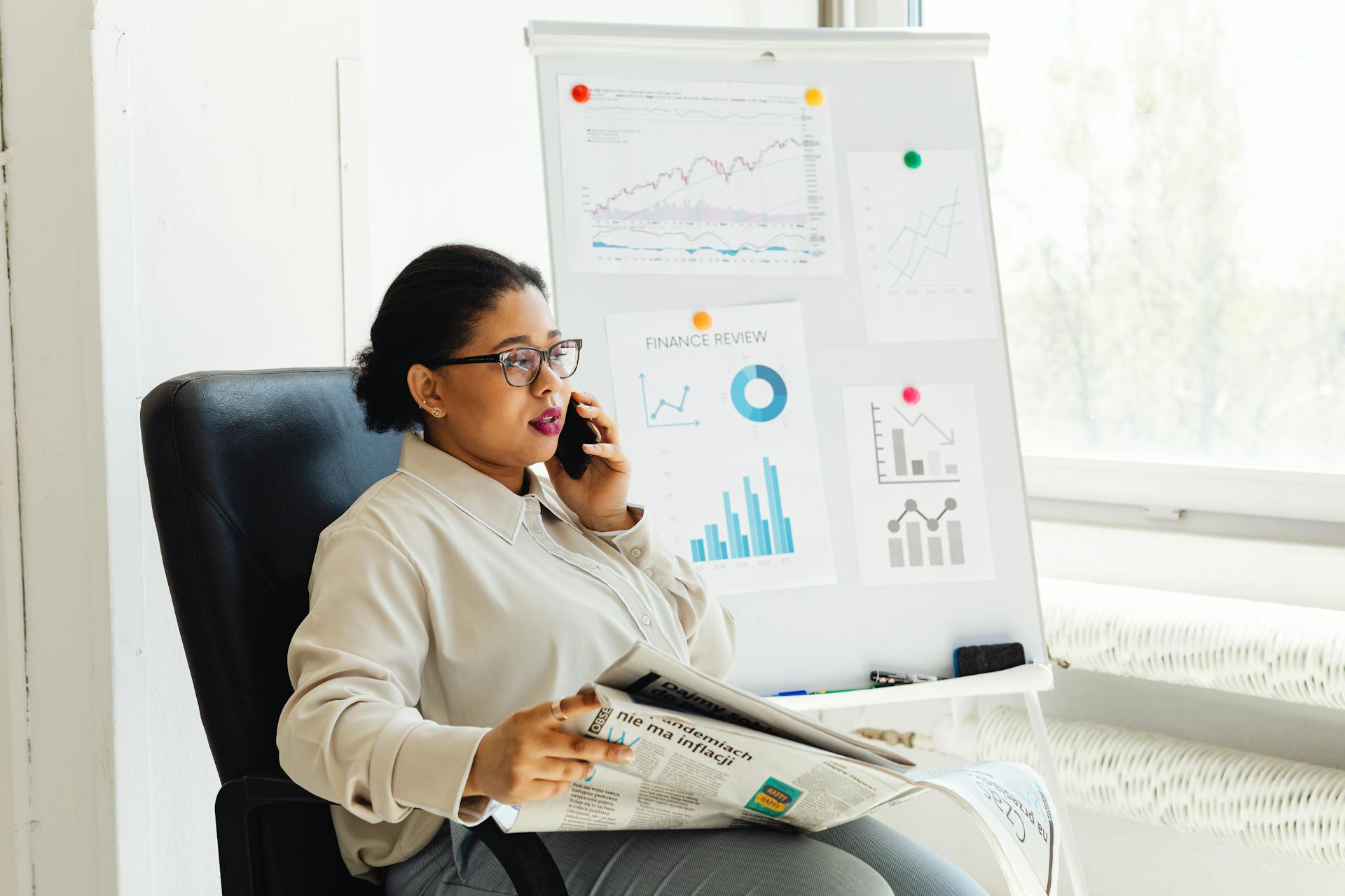 Businesswoman multitasking with phone and newspaper in office, examining financial charts.