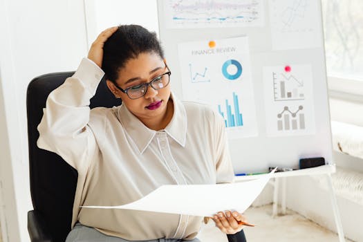 An adult woman examines financial reports at her office desk with charts and graphs in the background.