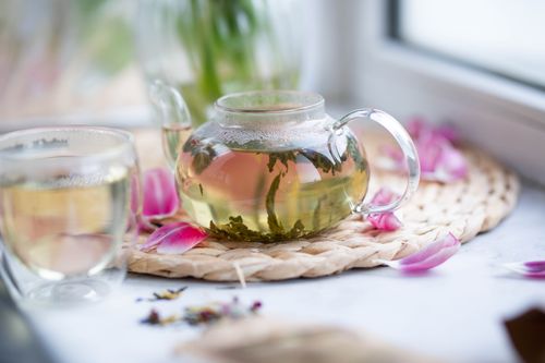 Free Transparent teapot with loose leaf tea placed on wicker mat with petals on white windowsill with glass on blurred background Stock Photo
