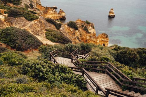 Free Wooden stairs lead to stunning Algarve cliffs with lush greenery and turquoise waters. Stock Photo