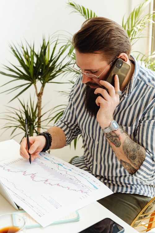Free A bearded man talking on a smartphone while examining financial charts with a pen in an office setting. Stock Photo